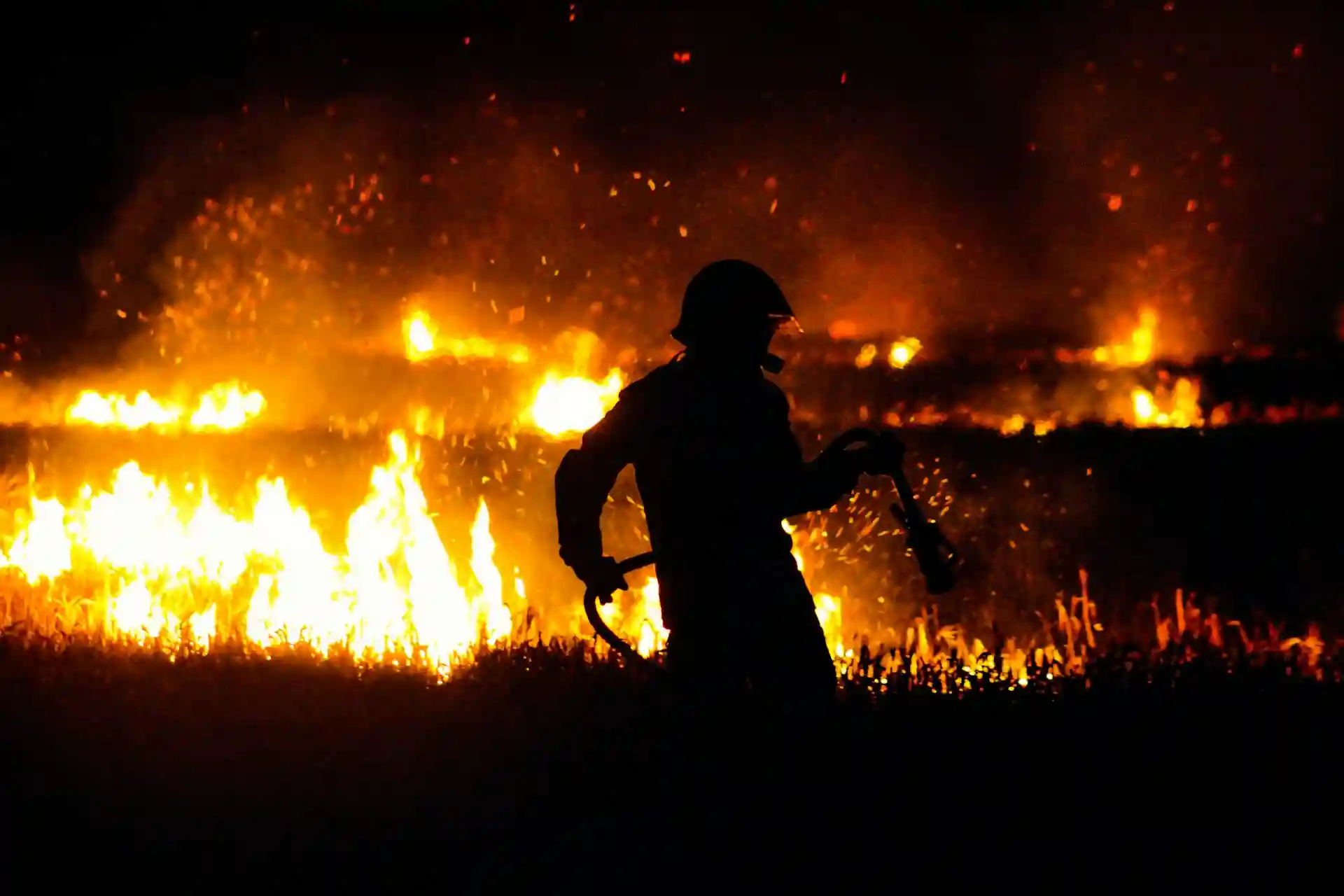 A firefighter silhouetted against a raging fire at night, holding a hose and navigating through grass as flames blaze in the background.