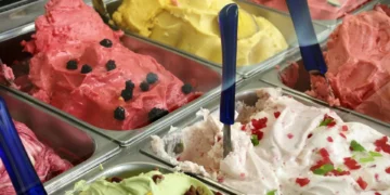 An assortment of colorful ice cream and gelato flavors displayed in a freezer, each with a blue-handled scooper, brings delight to customers but always recall to check for Listeria warnings for safety.