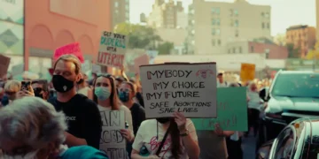 A crowd of people in a city street are holding signs, with one prominent sign reading "MY BODY MY CHOICE MY FUTURE SAFE ABORTIONS SAVE LIVES." Some individuals are wearing masks, possibly influenced by recent rulings like the Idaho abortion case heard by the U.S. Supreme Court.