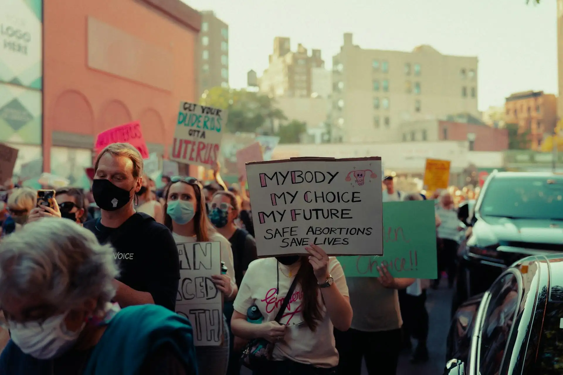 A crowd of people in a city street are holding signs, with one prominent sign reading "MY BODY MY CHOICE MY FUTURE SAFE ABORTIONS SAVE LIVES." Some individuals are wearing masks, possibly influenced by recent rulings like the Idaho abortion case heard by the U.S. Supreme Court.