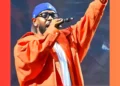 A person wearing an orange jacket, blue cap, and glasses, holding a microphone and raising one hand, performing on stage at an LA concert.