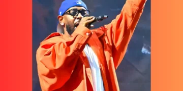 A person wearing an orange jacket, blue cap, and glasses, holding a microphone and raising one hand, performing on stage at an LA concert.