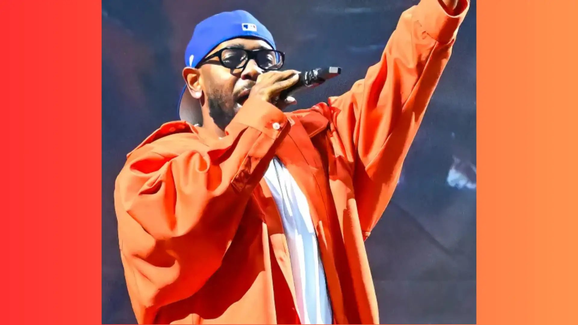 A person wearing an orange jacket, blue cap, and glasses, holding a microphone and raising one hand, performing on stage at an LA concert.