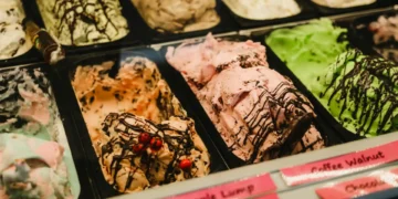Close-up of various ice cream pints in a display, including chocolate, mint, and strawberry, some with toppings like chocolate drizzles and small candies. Labels are partially visible at the bottom. Don't miss out on the Kroger ice cream giveaway for a chance to enjoy these delightful flavors for free!