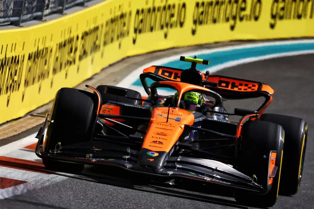 A Formula 1 car with an orange and black livery is speeding on a race track during the F1 Spanish GP 2024, surrounded by yellow advertising banners. The driver, potentially Lando Norris aiming for pole position, is focused, and the background includes a crowd in the stands enjoying the Spanish Grand Prix highlights.