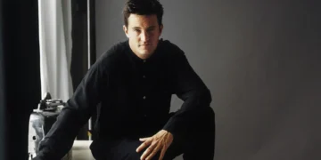 A person in dark clothing sits on a stool with one knee up, looking at the camera against a plain background, reminiscent of Matthew Perry's reflective moments.