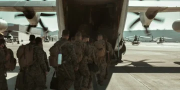 Military personnel in camouflage uniforms, including men aged 18-26, board a large aircraft with spinning propellers on the tarmac.