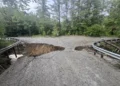 A rural road in New England is severely damaged by erosion, with a large section missing. Guardrails on both sides remain intact, and dense trees are visible in the background.