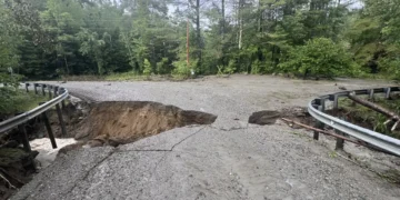 A rural road in New England is severely damaged by erosion, with a large section missing. Guardrails on both sides remain intact, and dense trees are visible in the background.