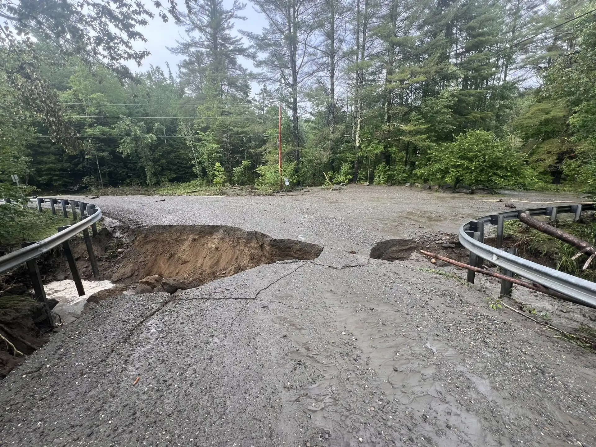 A rural road in New England is severely damaged by erosion, with a large section missing. Guardrails on both sides remain intact, and dense trees are visible in the background.