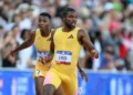 Two athletes in yellow uniforms run on a track during the U.S. Olympic Trials. The athlete in the foreground appears to be passing a baton to the athlete in the background during a relay race. In this moment, Noah Lyles showcases his speed and skill as they compete in the 200m event. A crowd is visible in the background.