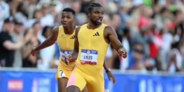 Two athletes in yellow uniforms run on a track during the U.S. Olympic Trials. The athlete in the foreground appears to be passing a baton to the athlete in the background during a relay race. In this moment, Noah Lyles showcases his speed and skill as they compete in the 200m event. A crowd is visible in the background.