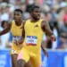 Two athletes in yellow uniforms run on a track during the U.S. Olympic Trials. The athlete in the foreground appears to be passing a baton to the athlete in the background during a relay race. In this moment, Noah Lyles showcases his speed and skill as they compete in the 200m event. A crowd is visible in the background.