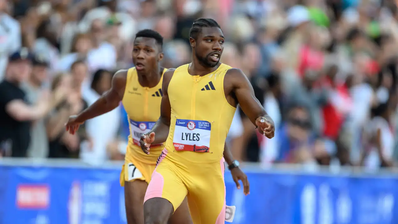 Two athletes in yellow uniforms run on a track during the U.S. Olympic Trials. The athlete in the foreground appears to be passing a baton to the athlete in the background during a relay race. In this moment, Noah Lyles showcases his speed and skill as they compete in the 200m event. A crowd is visible in the background.