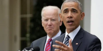 Barack Obama and Joe Biden in formal attire stand behind a podium outdoors. One is speaking into the microphone, while the other is looking on, reminiscent of an Obama-Biden debate moment.
