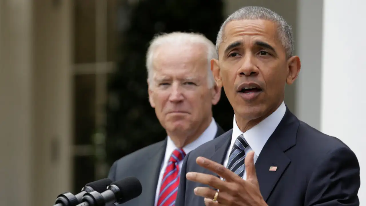 Barack Obama and Joe Biden in formal attire stand behind a podium outdoors. One is speaking into the microphone, while the other is looking on, reminiscent of an Obama-Biden debate moment.