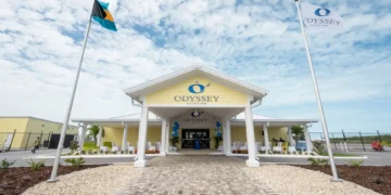 A yellow building with white columns labeled "Odyssey Aviation Exuma," flanked by two flagpoles, one with the Bahamian flag and one with the "Odyssey Aviation" flag, stands proudly under a partly cloudy sky, illustrating the charm of Caribbean infrastructure.