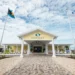 A yellow building with white columns labeled "Odyssey Aviation Exuma," flanked by two flagpoles, one with the Bahamian flag and one with the "Odyssey Aviation" flag, stands proudly under a partly cloudy sky, illustrating the charm of Caribbean infrastructure.