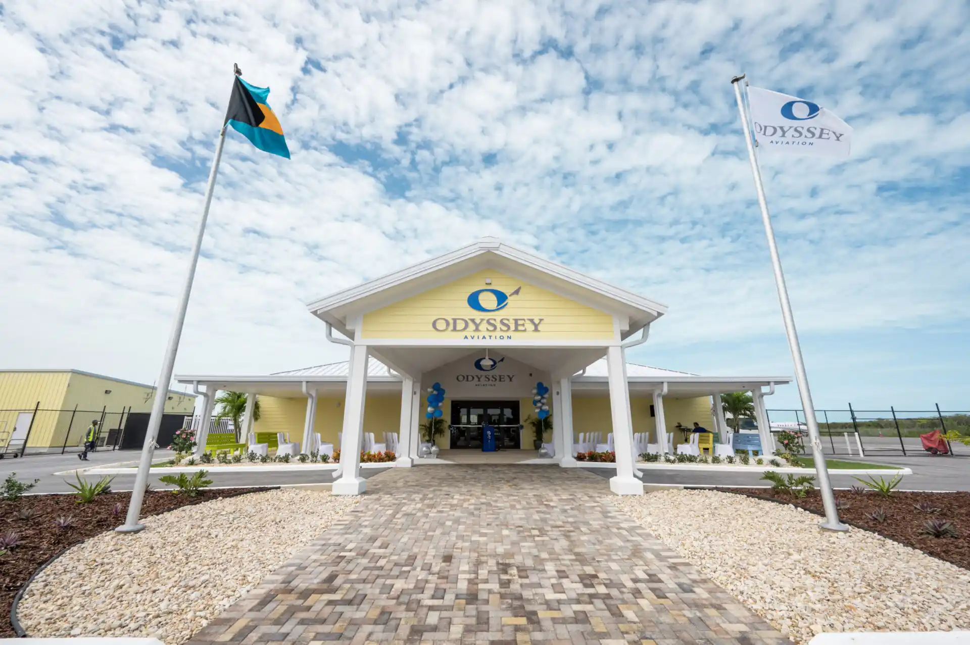 A yellow building with white columns labeled "Odyssey Aviation Exuma," flanked by two flagpoles, one with the Bahamian flag and one with the "Odyssey Aviation" flag, stands proudly under a partly cloudy sky, illustrating the charm of Caribbean infrastructure.
