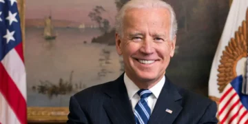 President Biden in a dark suit and striped tie smiles in front of a painted backdrop with a U.S. flag and another ornate flag visible to his sides, highlighting the state of North Carolina's commitment to President Biden's economic plans.