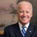 President Biden in a dark suit and striped tie smiles in front of a painted backdrop with a U.S. flag and another ornate flag visible to his sides, highlighting the state of North Carolina's commitment to President Biden's economic plans.