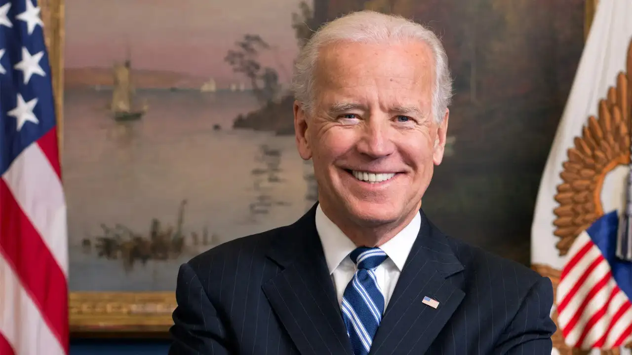 President Biden in a dark suit and striped tie smiles in front of a painted backdrop with a U.S. flag and another ornate flag visible to his sides, highlighting the state of North Carolina's commitment to President Biden's economic plans.