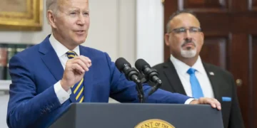 A man in a blue suit speaks at a podium with the presidential seal, addressing the Israel-Hamas ceasefire, while another man in a suit and tie stands behind him in a room with a U.S. flag and bookshelf.