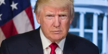 A person with blonde hair and a suit sits in front of an American flag and a blurred building background, reminiscent of recent appearances involving Trump amidst his legal challenges.