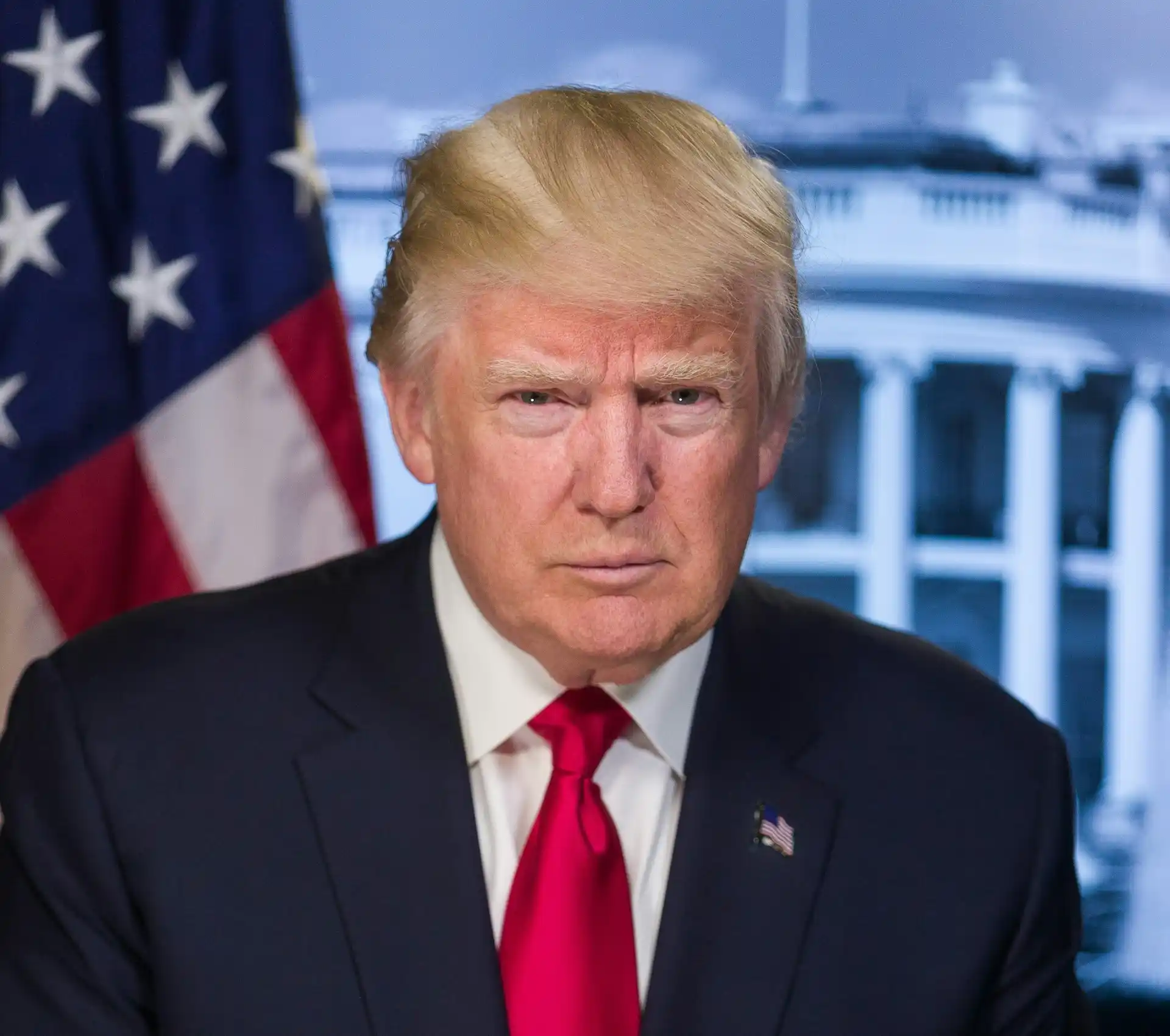 A person with blonde hair and a suit sits in front of an American flag and a blurred building background, reminiscent of recent appearances involving Trump amidst his legal challenges.