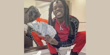 Julio Foolio, dressed in a red and black tracksuit, is squatting on a bathroom counter, holding a fan of $100 bills and smiling, with his reflection visible in the mirror behind.