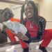 Julio Foolio, dressed in a red and black tracksuit, is squatting on a bathroom counter, holding a fan of $100 bills and smiling, with his reflection visible in the mirror behind.