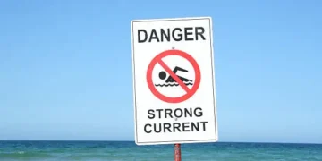 A sign on a Florida beach reads "Danger Strong Current" with an icon of a swimmer inside a red circle with a diagonal line through it, warning visitors of dangerous rip currents. The ocean and waves are in the background, emphasizing the risk of drownings if one ignores the caution.