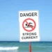 A sign on a Florida beach reads "Danger Strong Current" with an icon of a swimmer inside a red circle with a diagonal line through it, warning visitors of dangerous rip currents. The ocean and waves are in the background, emphasizing the risk of drownings if one ignores the caution.