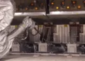 A robotic arm operates in an industrial setting at Pixelle Specialty Solutions, manipulating a row of switches on a complex machinery panel, with multiple lit indicators in the background. The system enhances efficiency, particularly in tasks like recovery boiler operation and smelt spout cleaning.