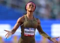 ShaCarri-Richardson with a "Richardson" name tag stands on a track with arms outstretched, basking in the Olympic glory, wearing a gold-toned uniform and a pink headband.