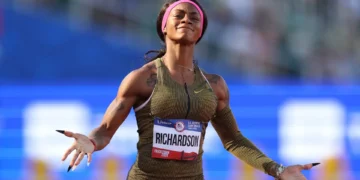ShaCarri-Richardson with a "Richardson" name tag stands on a track with arms outstretched, basking in the Olympic glory, wearing a gold-toned uniform and a pink headband.