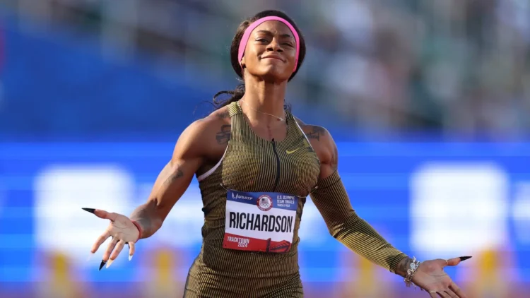 ShaCarri-Richardson with a "Richardson" name tag stands on a track with arms outstretched, basking in the Olympic glory, wearing a gold-toned uniform and a pink headband.