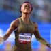 ShaCarri-Richardson with a "Richardson" name tag stands on a track with arms outstretched, basking in the Olympic glory, wearing a gold-toned uniform and a pink headband.