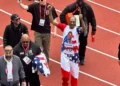 A man wearing a patriotic outfit with a graphic of Snoop Dogg on it is walking on the track, smiling, and giving thumbs up during the Olympic Trials 200m event. He is surrounded by several other people.