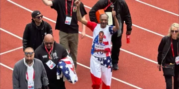 A man wearing a patriotic outfit with a graphic of Snoop Dogg on it is walking on the track, smiling, and giving thumbs up during the Olympic Trials 200m event. He is surrounded by several other people.