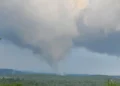 A tornado funnel cloud touches down in a rural Connecticut landscape with forested hills under a cloudy sky, causing widespread damage.