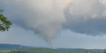 A tornado funnel cloud touches down in a rural Connecticut landscape with forested hills under a cloudy sky, causing widespread damage.