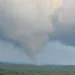 A tornado funnel cloud touches down in a rural Connecticut landscape with forested hills under a cloudy sky, causing widespread damage.