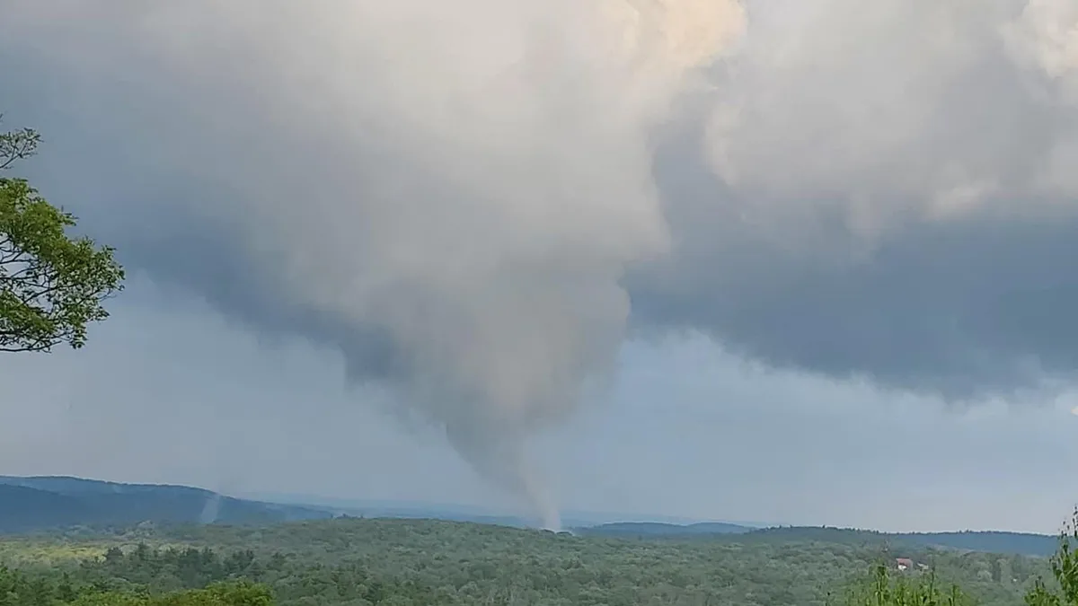 A tornado funnel cloud touches down in a rural Connecticut landscape with forested hills under a cloudy sky, causing widespread damage.