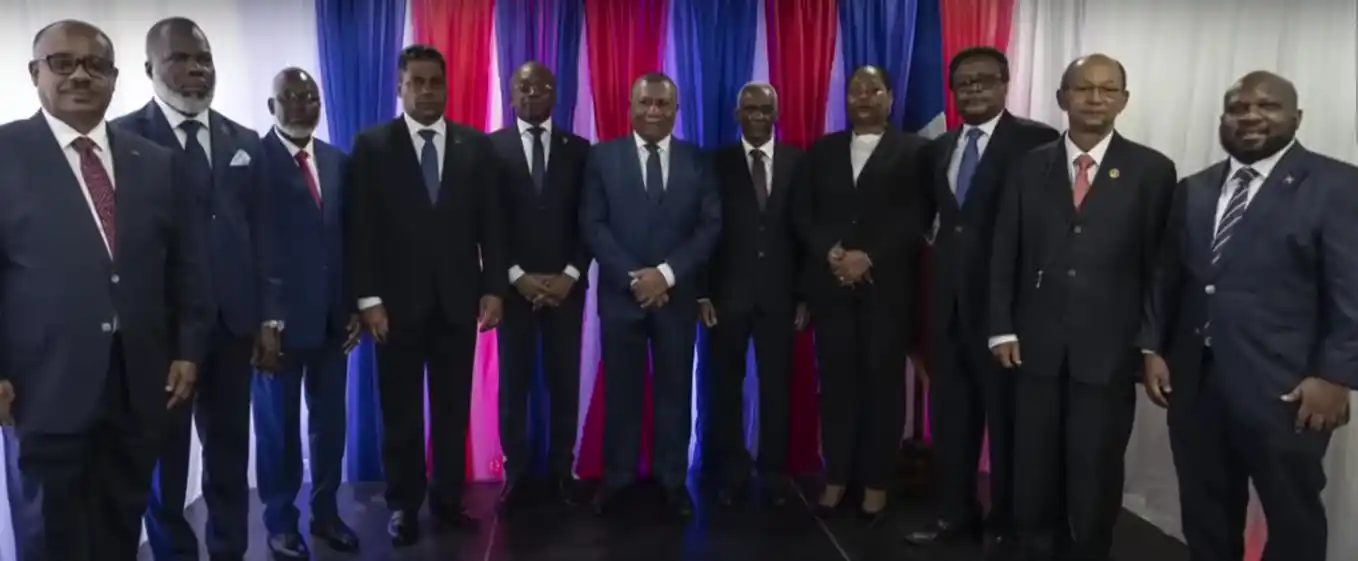 A group of eleven people in formal attire stands in front of red, white, and blue striped curtains, posing for a photo to mark Haiti's new government formation.