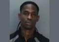 A man with long braids and a serious expression is seen against a plain background, wearing a dark shirt with light gray accents, resembling the stern ambiance of the Miami Beach Marina.