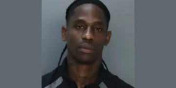 A man with long braids and a serious expression is seen against a plain background, wearing a dark shirt with light gray accents, resembling the stern ambiance of the Miami Beach Marina.