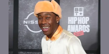 Person smiling and wearing an orange hat and light-colored shirt, standing in front of a backdrop with "BET Hip Hop Awards 2021," "Nissan" logos, reminiscent of the vibrant energy seen at Lollapalooza.