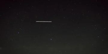 A long-exposure photograph of the night sky above Red Rocks Amphitheatre shows a streak of light, likely from a satellite or a meteor, among numerous visible stars.