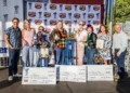 A group stands on a stage holding trophies and large checks in front of a backdrop with Sonoma Marin Fair logos. Four people are holding certificates, celebrating the World's Ugliest Dog 2024 event, with others standing alongside them.